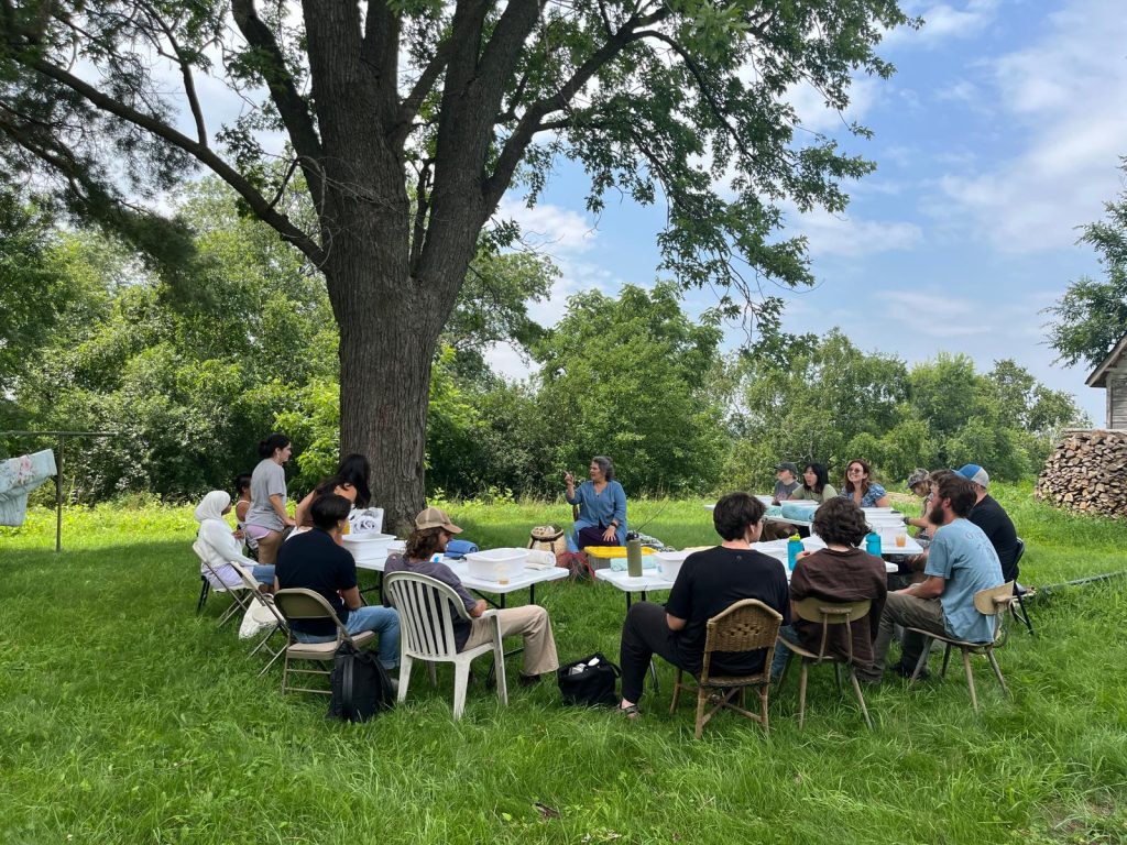A dozen people sitting around a table under a tree listening to someone presenting